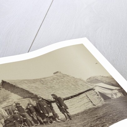 A group of soldiers, and two young men, one an African American, stand outside of log cabin quarters by American Photographer