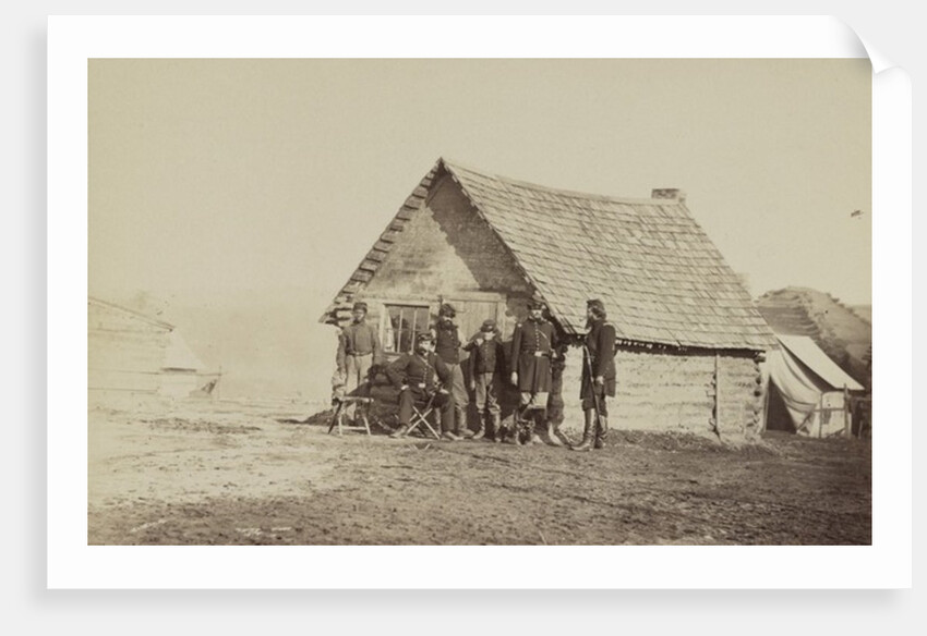 A group of soldiers, and two young men, one an African American, stand outside of log cabin quarters by American Photographer