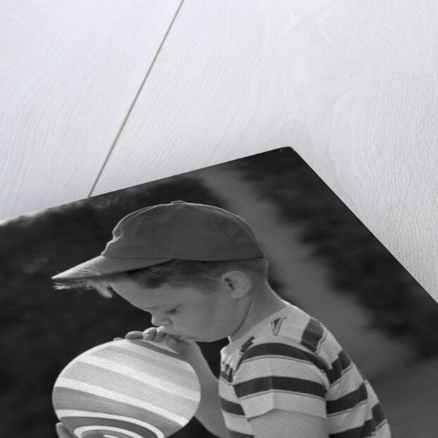 1950s Side View Of Boy In Baseball Cap and Striped T-Shirt Blowing Up Swirled Balloon by Anonymous
