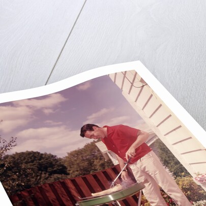 1960s Man Wearing Red Shirt Cooking Steak Outdoor On Backyard Grill by Anonymous