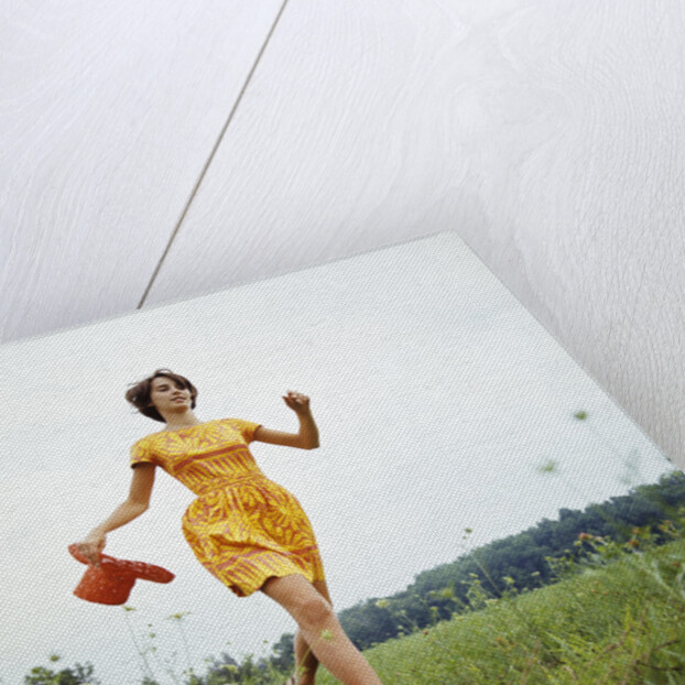 1970s Young Woman Wearing Colorful Print Dress Running In Field Carrying Red Straw Hat by Anonymous