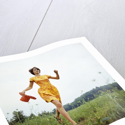 1970s Young Woman Wearing Colorful Print Dress Running In Field Carrying Red Straw Hat by Anonymous