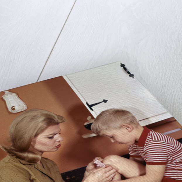 1970s Mother Giving First-Aid Putting Band-Aid On Knee Of Son In Kitchen by Anonymous