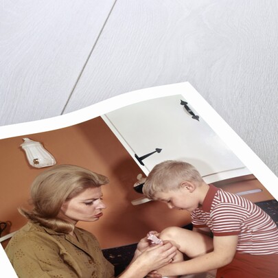 1970s Mother Giving First-Aid Putting Band-Aid On Knee Of Son In Kitchen by Anonymous