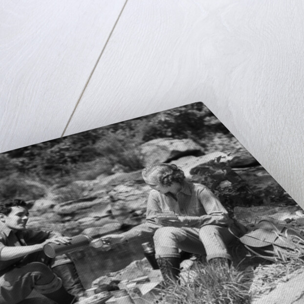 Couple having picnic sitting on rocks by stream man pouring drink for woman from thermos by Anonymous