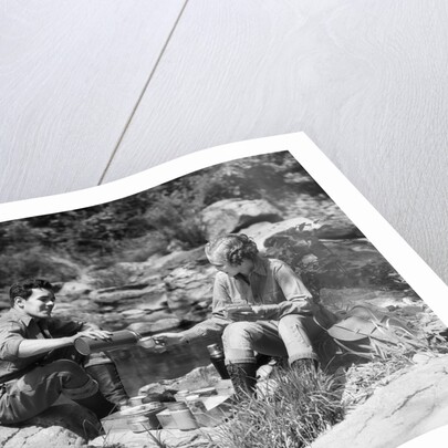 Couple having picnic sitting on rocks by stream man pouring drink for woman from thermos by Anonymous