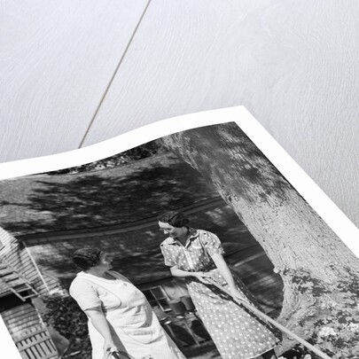 Two women talking in yard one holding garden hoe the other holds basket with flowers by Anonymous