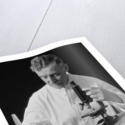 Male scientist wearing white lab coat writing data on clip board holding focus control of microscope by Anonymous