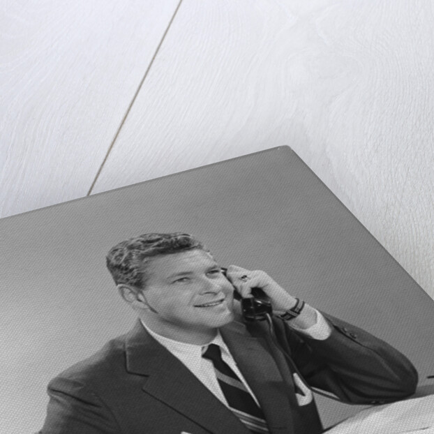 Businessman seated at desk talking on telephone smiling writing with pencil by Anonymous