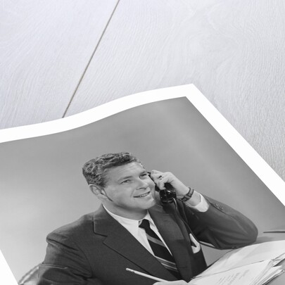 Businessman seated at desk talking on telephone smiling writing with pencil by Anonymous