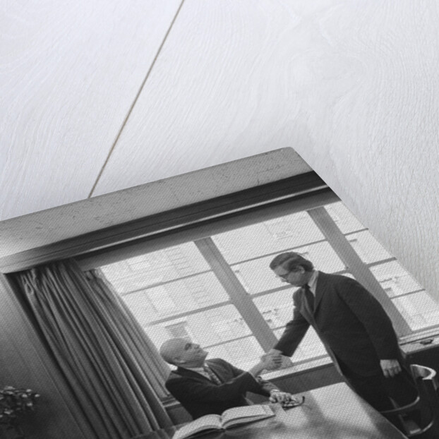 Two men one seated shaking hands long wooden conference table by Anonymous