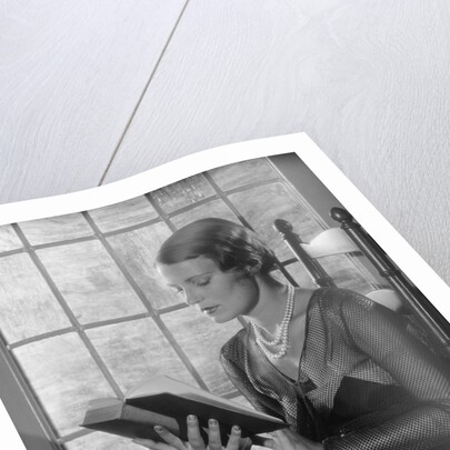 Elegant woman wearing pearls sitting by window reading book by Anonymous