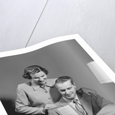 Couple man seated at desk holding paper woman standing looking over his shoulder smiling happy by Anonymous