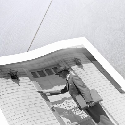 Salesman wearing suit tie hat carrying briefcase ringing suburban house doorbell by Anonymous