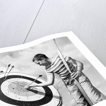 1960s boy jeans striped t-shirt holding bow & pulling arrow out of target bull's-eye by Anonymous