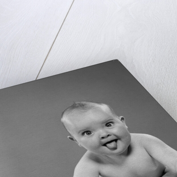 1950s portrait of baby sitting and sticking tongue out looking at camera by Anonymous