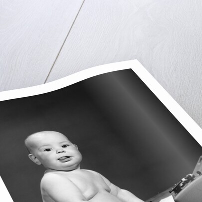 1950s 1960s baby in diaper sticking out tongue holding glasses sitting before adding machine looking at camera by Anonymous