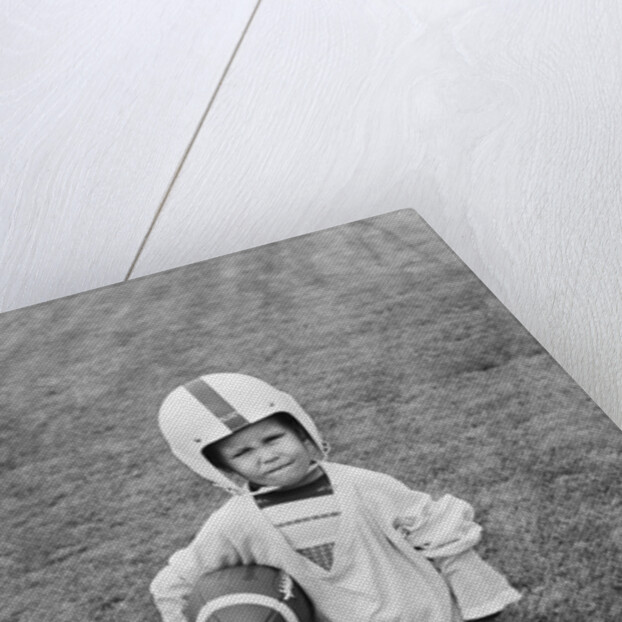 1950s boy standing in grass wearing oversized shirt & helmet holding football by Anonymous