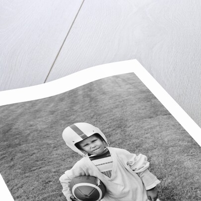 1950s boy standing in grass wearing oversized shirt & helmet holding football by Anonymous