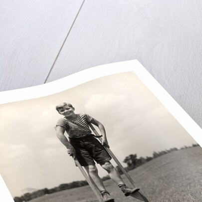 1930s 1940s smiling happy boy wearing striped shirt & short pants walking on pair of stilts looking at camera by Anonymous