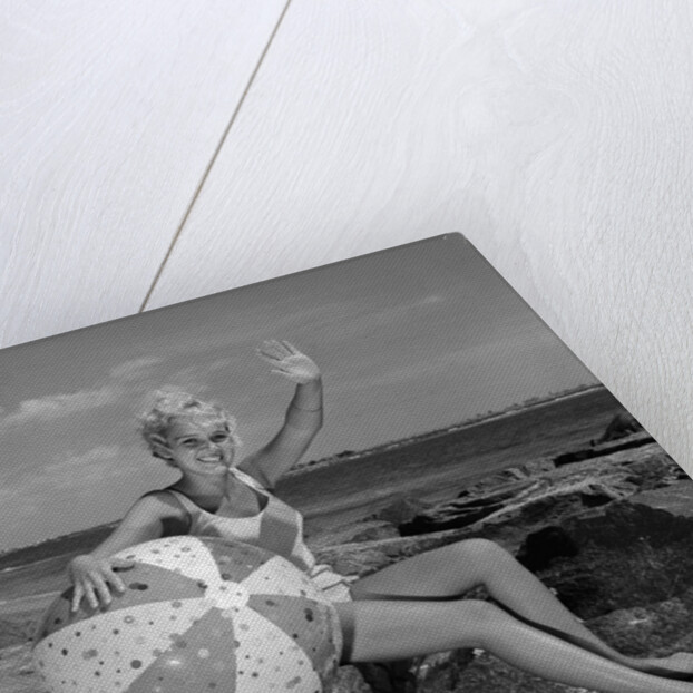 1960s smiling blond teenage girl sitting on rocky shore holding a beach ball waving looking at camera by Anonymous