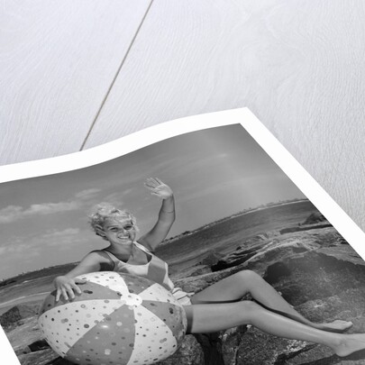 1960s smiling blond teenage girl sitting on rocky shore holding a beach ball waving looking at camera by Anonymous