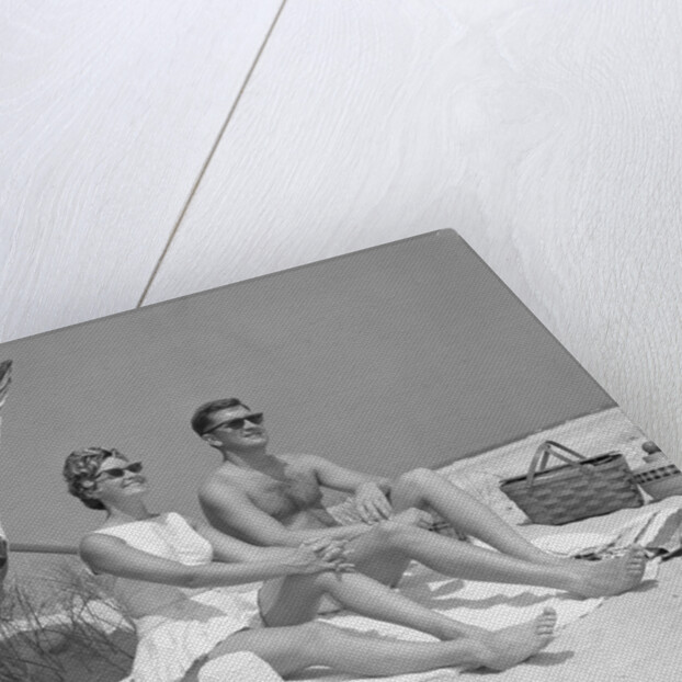 1960s couple in sunglasses sitting on beach blanket with legs extended with umbrella by Anonymous