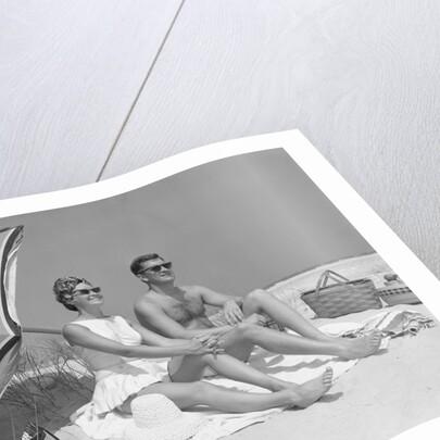 1960s couple in sunglasses sitting on beach blanket with legs extended with umbrella by Anonymous