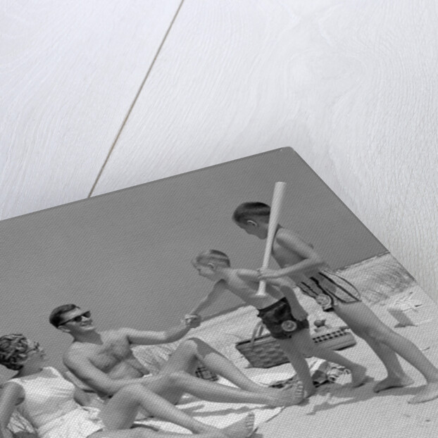 1960s at the beach two boys with baseball bat trying to pull up father seated next to mother so he will play with them by Anonymous