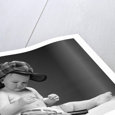 1960s baby in diaper wearing sherlock holmes deerstalker style double-billed cap with magnifying glass resting on leg inspecting a clue by Anonymous
