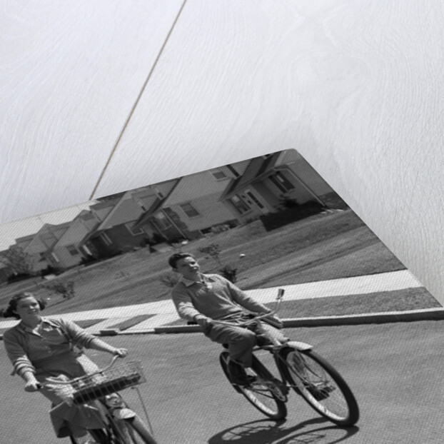 1950s teen boy girl couple riding bikes down residential street by Anonymous