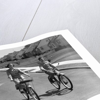 1950s teen boy girl couple riding bikes down residential street by Anonymous