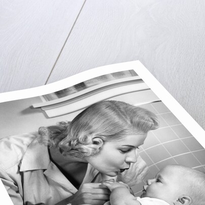 1950s mother woman bending over baby lying on layette holding hands talking by Anonymous