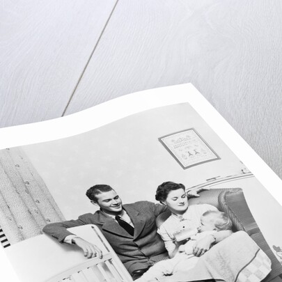 1930s 1940s mother sitting in chair in nursery baby 11 months on lap drinking from bottle father looking on leaning on crib by Anonymous