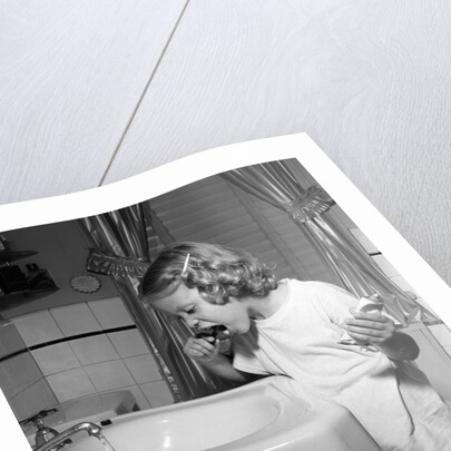 1950s little blond girl in pajamas standing at bathroom sink brushing her teeth toothpaste and toothbrush in hand by Anonymous