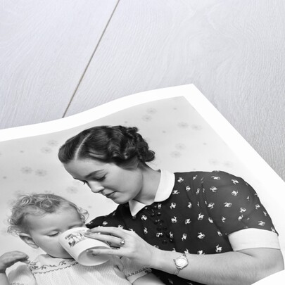 1930s woman mother giving baby daughter drink from mug by Anonymous