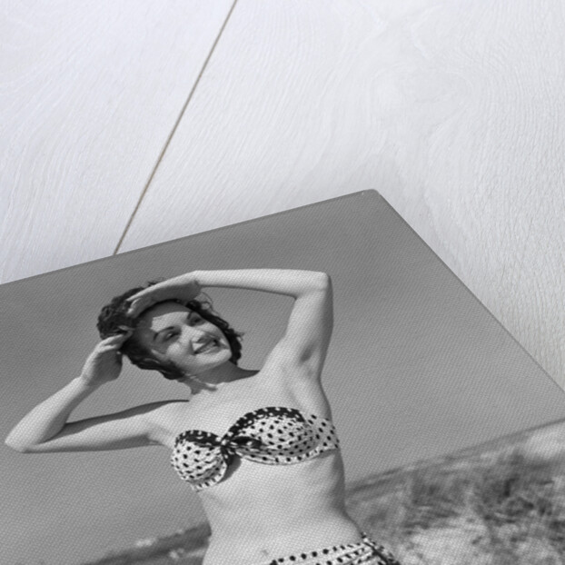 1950s smiling young woman kneeling in grassy sand wearing polka dot bikini shading eyes from sun by Anonymous