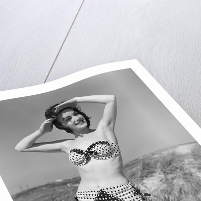 1950s smiling young woman kneeling in grassy sand wearing polka dot bikini shading eyes from sun by Anonymous