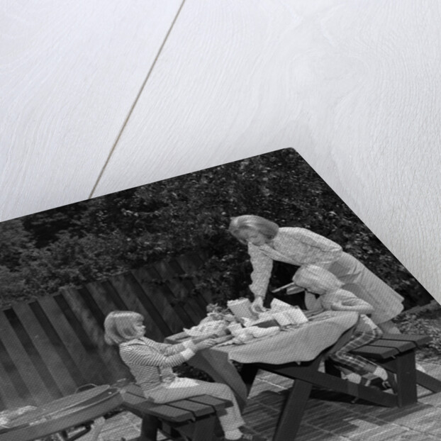 1960s man on patio grilling steak with 2 daughters seated at picnic table & wife standing serving food by Anonymous