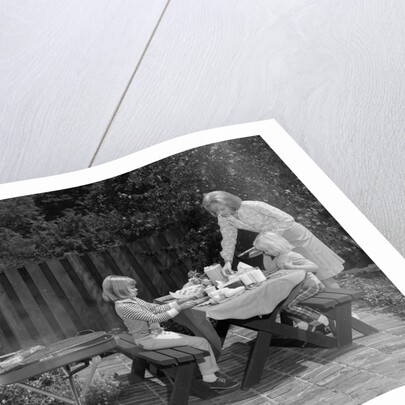 1960s man on patio grilling steak with 2 daughters seated at picnic table & wife standing serving food by Anonymous