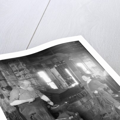 1930s elderly blacksmith with hammer at anvil as young boy holding harness looks on smiling by Anonymous