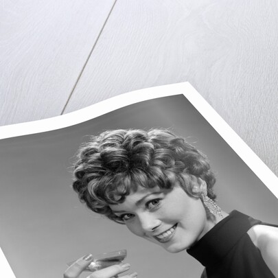 1960s glamorous woman with curly hair and dangling earrings smiling holding a champagne glass looking at camera by Anonymous