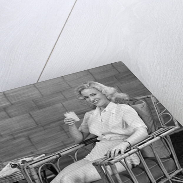 1940s blonde woman in bamboo chair holding up glass of milk smiling wearing white shorts looking at camera by Anonymous