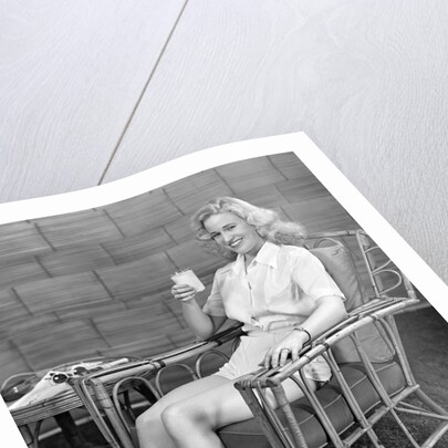 1940s blonde woman in bamboo chair holding up glass of milk smiling wearing white shorts looking at camera by Anonymous