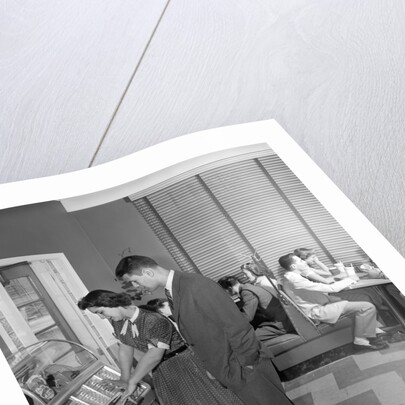 1950s teen couple playing juke box in malt shop with other teens in booths by Anonymous