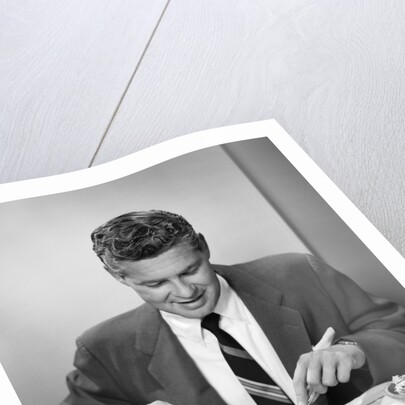 1950s smiling man in suit and tie sitting at table holding knife and fork eating dinner by Anonymous