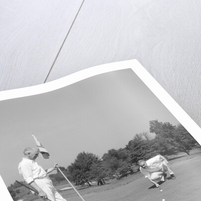1930s 1940s elderly men on golf green one holding flag the other kneels lining up his putt by Anonymous
