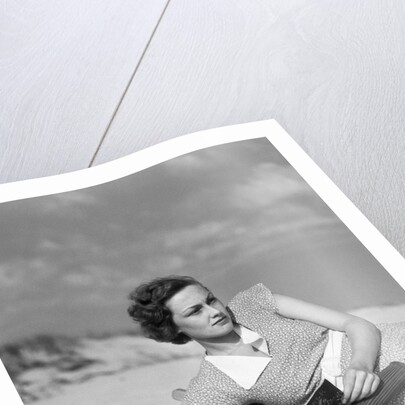 1930s woman leaning in wooden chair on beach wearing printed dress holding magazine looking thoughtful by Anonymous