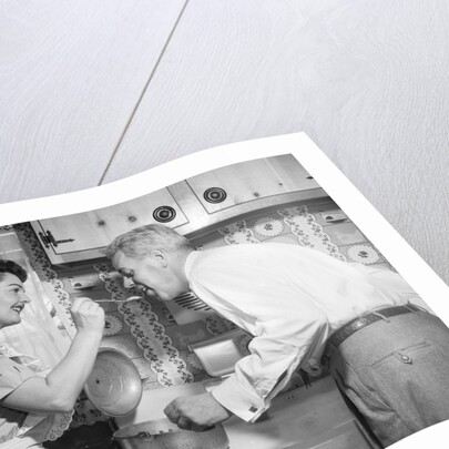 1950s housewife in kitchen having husband taste food on stove by Anonymous