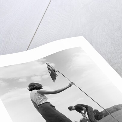 1970s couple playing golf man putting woman holding flag by Anonymous